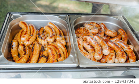 Juicy grilled sausages in two stainless steel buffet trays, ready to serve at outdoor catering events, parties, or barbecues. Perfect for food service, hospitality, and menu illustration. Juicy grilled sausages in two stainless steel buffet trays, ready to serve at outdoor catering events, parties, or barbecues. Perfect for food service, hospitality, and menu illustration. 129437342