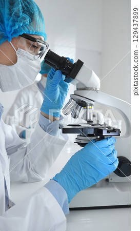 Female scientist wearing a mask and blue safety gear, using a microscope for research while a male colleague works in the background. Medicine, healthcare and science concept 129437899