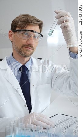 Laboratory professional examining a blue liquid test tube while documenting research in a protective coat, glasses, and gloves. Medicine, health care and science concept 129437938