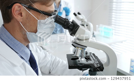 Medical researcher analyzing sample under microscope, wearing protective gear in sterile laboratory setting, performing scientific research. Medicine, healthcare and science concept 129437951
