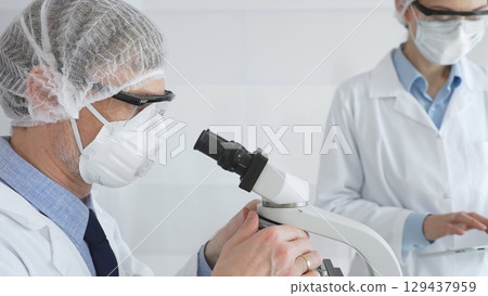 Senior male scientist wearing protective mask, glasses and hairnet using microscope with young female colleague assisting, holding tablet and taking notes in laboratory. Medicine and science concept Senior male scientist wearing protective mask, glasses and hairnet using microscope with young female colleague assisting, holding tablet and taking notes in laboratory. Medicine and science concept 129437959