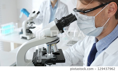 Masked scientist analyzing sample under microscope, colleague researching nearby in sterile medical laboratory setting. Medicine, healthcare and science concept Masked scientist analyzing sample under microscope, colleague researching nearby in sterile medical laboratory setting. Medicine, healthcare and science concept 129437962