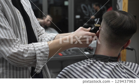 Professional barber grooming client's hair using electric clipper, delivering precise cut with skill in contemporary salon setting Professional barber grooming client's hair using electric clipper, delivering precise cut with skill in contemporary salon setting 129437963