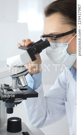 A female scientist in a protective mask and glasses carefully examines a sample under a microscope while conducting research in a modern laboratory. Concept of medicine and science A female scientist in a protective mask and glasses carefully examines a sample under a microscope while conducting research in a modern laboratory. Concept of medicine and science 129437967