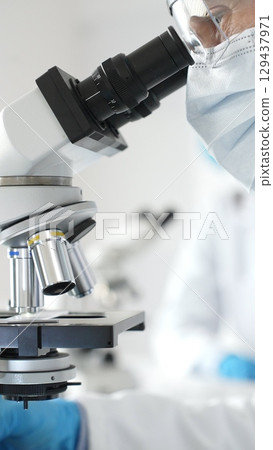 Close up of male researcher examining sample through microscope, wearing blue safety gear in modern laboratory setting. Medicine, healthcare and science concept Close up of male researcher examining sample through microscope, wearing blue safety gear in modern laboratory setting. Medicine, healthcare and science concept 129437971