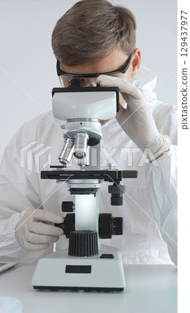 A male medical virologist in a medical gown and gloves examines viral samples under a microscope while conducting research in a sterile laboratory setting 129437977