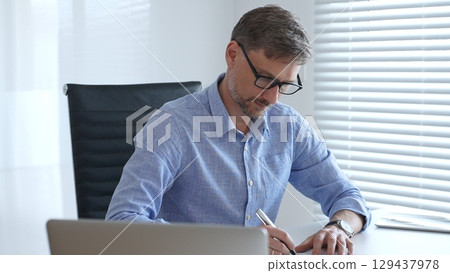 Male adult accountant wearing blue sleek shirt and black glasses is concentrating while working at the desk in modern office, demonstrating professionalism. Business people, taxes and audit concept 129437978