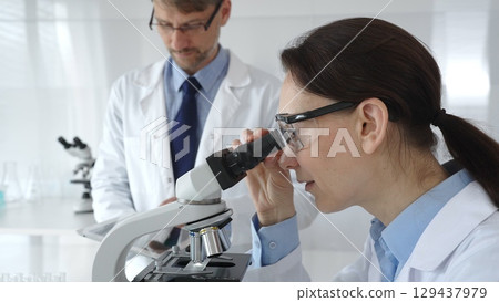 A female scientist wearing protective glasses looks into a microscope and conducts a medical research in a sterile laboratory while her male colleague works in the background. Medicine and science A female scientist wearing protective glasses looks into a microscope and conducts a medical research in a sterile laboratory while her male colleague works in the background. Medicine and science 129437979