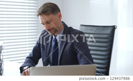Adult businessman wearing blue suit and tie. is sitting at a modern desk, working intently on a laptop surrounded by folders, showcasing professionalism and determination. Audit and taxes in business Adult businessman wearing blue suit and tie. is sitting at a modern desk, working intently on a laptop surrounded by folders, showcasing professionalism and determination. Audit and taxes in business 129437986