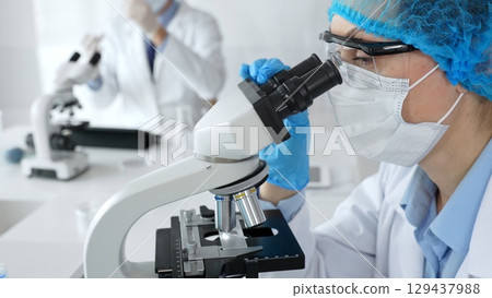 Female scientist wearing a mask and blue safety gear, using a microscope for research while a male colleague works in the background. Medicine, healthcare and science concept 129437988