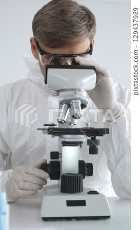 A male medical virologist in a medical gown and gloves examines viral samples under a microscope while conducting research in a sterile laboratory setting 129437989