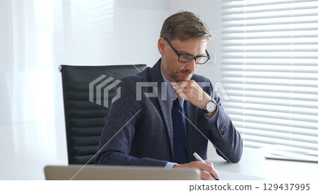 Adult businessman wearing blue suit and tail is concentrating and thinking while working at the desk in a bright, modern office, demonstrating professionalism and efficiency. Business people concept 129437995