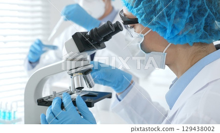 Close up of woman scientist wearing blue protective gear while examining specimen under microscope, laboratory assistant handles glassware in medical investigation at the background. Medicine concept 129438002