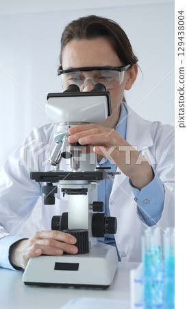 Female scientist wearing lab coat and protective glasses using microscope, analyzing samples in test tubes in modern laboratory, conducting scientific research 129438004