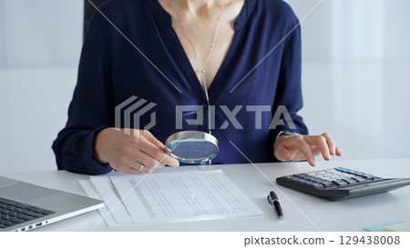 Professional female analyzing financial documents, using magnifying glass and calculator at workplace desk, focused on detailed accounting review, close up. Audit and taxes in business 129438008