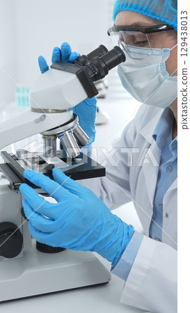 Female scientist analyzing sample through microscope, wearing protective lab gear during medical research investigation. Medicine, laboratory and science concept Female scientist analyzing sample through microscope, wearing protective lab gear during medical research investigation. Medicine, laboratory and science concept 129438013