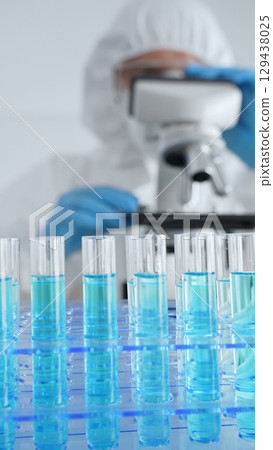 Close up of test tubes with blue liquid opposite a scientific researcher wearing protective gear tuning microscope lenses in laboratory. Medicine, healthcare and science concept Close up of test tubes with blue liquid opposite a scientific researcher wearing protective gear tuning microscope lenses in laboratory. Medicine, healthcare and science concept 129438025