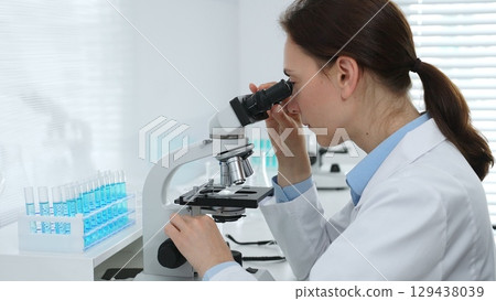 Female scientist examining microscope slide, analyzing blue chemical sample in sterile laboratory environment, performing precision medical research. Medicine, healthcare and science concept Female scientist examining microscope slide, analyzing blue chemical sample in sterile laboratory environment, performing precision medical research. Medicine, healthcare and science concept 129438039