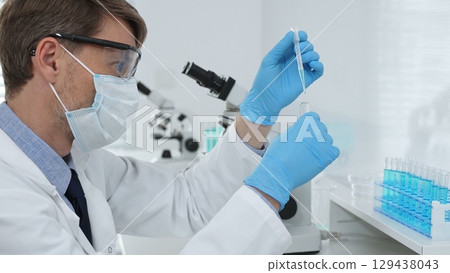 Masked male scientist in lab coat, protective glasses and blue gloves is adding reagent to test tube with microscopes and test tube rack in modern laboratory. Medicine, healthcare and science concept Masked male scientist in lab coat, protective glasses and blue gloves is adding reagent to test tube with microscopes and test tube rack in modern laboratory. Medicine, healthcare and science concept 129438043