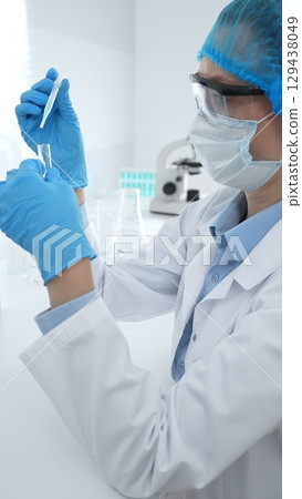 Medical science researcher wearing blue protective gear of cap, gloves and googles, is examining liquid in test tube in laboratory workspace. Medicine and health care concept 129438049