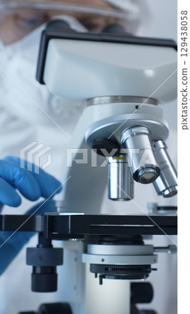 Close up of medical virologist in a hazmat suit, mask, and gloves, diligently working at a microscope in a laboratory researching viruses. Medicine and science concept Close up of medical virologist in a hazmat suit, mask, and gloves, diligently working at a microscope in a laboratory researching viruses. Medicine and science concept 129438058