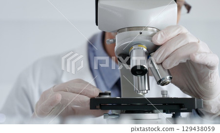 Close up of scientific researcher wearing white protective gloves carefully calibrating laboratory microscope, performing precision medical investigation. Medicine and science concept 129438059