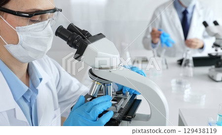Female scientist wearing mask and gloves using microscope doing scientific research in modern laboratory with colleague working in background. Medicine, healthcare and science concept 129438119