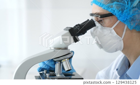 Female scientist examining sample through microscope, wearing protective lab gear while conducting medical research. Laboratory and science concept 129438125