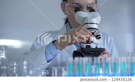 A female scientist, wearing a lab coat and safety glasses, is adjusting a binocular microscope surrounded by test tubes in laboratory setting. Medicine and science 129438126