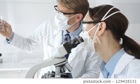 Two scientists, a man and a woman, in lab coats and protective gear, conduct research in a sterile laboratory. Concept of medicine and science Two scientists, a man and a woman, in lab coats and protective gear, conduct research in a sterile laboratory. Concept of medicine and science 129438131