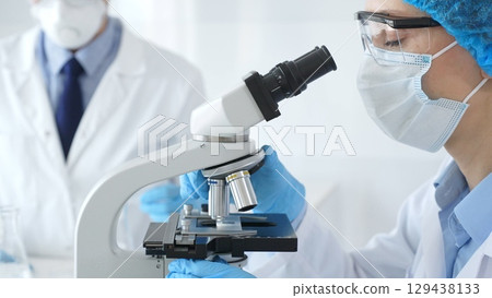 Close up of woman scientist wearing blue protective gear while examining specimen under microscope, laboratory assistant handles glassware in medical investigation at the background. Medicine concept Close up of woman scientist wearing blue protective gear while examining specimen under microscope, laboratory assistant handles glassware in medical investigation at the background. Medicine concept 129438133