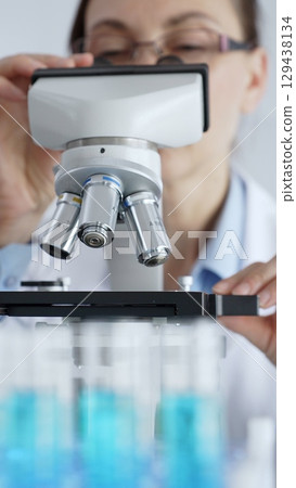Female scientist wearing protective equipment checking microscope, analyzing blue test tubes in modern laboratory research setting. Medicine, healthcare and science concept Female scientist wearing protective equipment checking microscope, analyzing blue test tubes in modern laboratory research setting. Medicine, healthcare and science concept 129438134