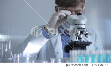 Male scientist, wearing a lab coat, mask, white gloves and safety glasses, is using a binocular microscope surrounded by test tubes in laboratory Male scientist, wearing a lab coat, mask, white gloves and safety glasses, is using a binocular microscope surrounded by test tubes in laboratory 129438135