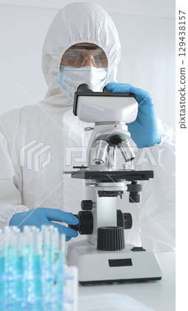 A medical virologist in protective gear examines viral samples under a microscope while conducting research in a laboratory setting 129438157