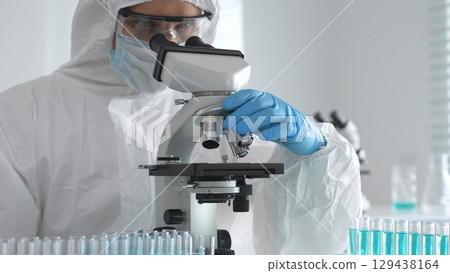 Medical virologist in protective hazmat gear examining viral samples under a microscope while conducting research in a sterile laboratory environment Medical virologist in protective hazmat gear examining viral samples under a microscope while conducting research in a sterile laboratory environment 129438164