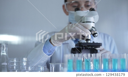 A scientist woman, wearing a lab coat, mask, white gloves and safety glasses, is adjusting a microscope surrounded by test tubes in laboratory 129438179