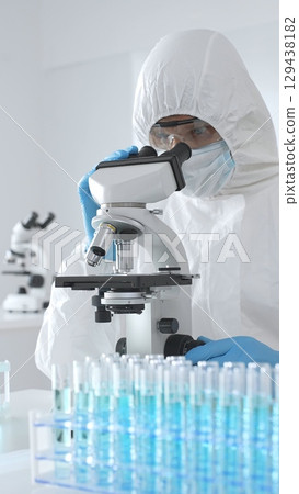Medical scientist in protective suit using microscope while analyzing samples, conducting research in sterile laboratory environment. Close-up of test tubes with blue liquid Medical scientist in protective suit using microscope while analyzing samples, conducting research in sterile laboratory environment. Close-up of test tubes with blue liquid 129438182