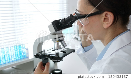 Female scientist examining sample under microscope, conducting research in laboratory with colleague working near test tube rack. Concept of medicine, healthcare and science Female scientist examining sample under microscope, conducting research in laboratory with colleague working near test tube rack. Concept of medicine, healthcare and science 129438187