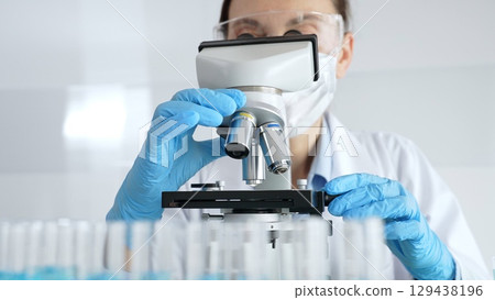 Woman scientist wearing protective gear is checking microscope, analyzing blue test tubes in modern laboratory research setting. Medicine, healthcare and science concept 129438196