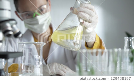 Woman scientist researcher wearing white protective gloves is shaking up a yellow chemical solution inside an Erlenmeyer flask in a laboratory setting, portrait view. Science and medicine Woman scientist researcher wearing white protective gloves is shaking up a yellow chemical solution inside an Erlenmeyer flask in a laboratory setting, portrait view. Science and medicine 129438197