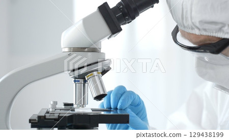 Medical virologist in a hazmat suit, mask, and blue gloves, diligently working at a microscope in a laboratory researching viruses. Healthcare, medicine, and science concept 129438199