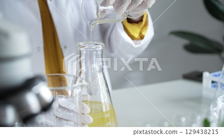 A scientist wearing a lab coat and white gloves is pouring a yellow oily liquid from one beaker to another in a laboratory setting, close up. Science and medicine A scientist wearing a lab coat and white gloves is pouring a yellow oily liquid from one beaker to another in a laboratory setting, close up. Science and medicine 129438215
