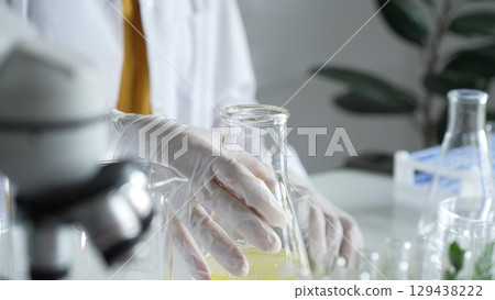 A scientist wearing white protective gloves is shaking up a yellow chemical solution inside an Erlenmeyer flask in a laboratory set, close up. Science and medicine concepts 129438222