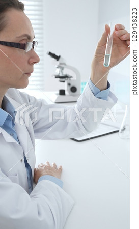 Female researcher examining clear liquid in test tube, sitting near microscopes and lab equipment within sterile scientific workspace. Medicine, healthcare and science concept 129438223