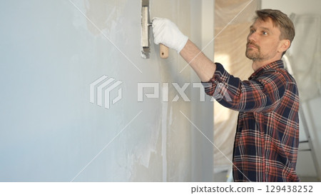 Male construction worker wearing gloves is carefully applying plaster to a wall using a hand tool, contributing to a home renovation or construction project 129438252