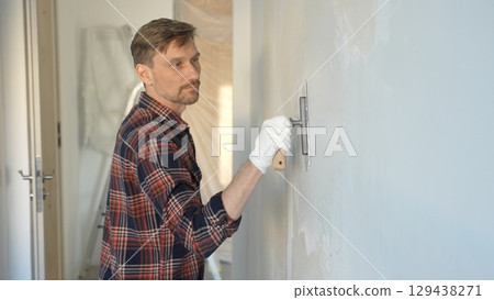 Male construction worker wearing gloves is carefully applying plaster to a wall using a hand tool, contributing to a home renovation or construction project 129438271