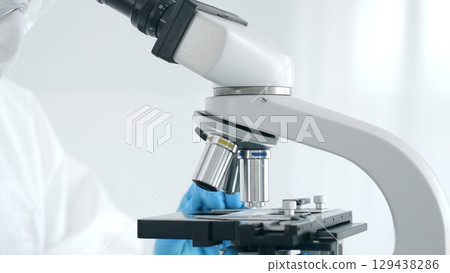 Close up of medical virologist in a hazmat suit, mask, blue gloves, working at a microscope in a laboratory researching viruses. Healthcare and science concept 129438286