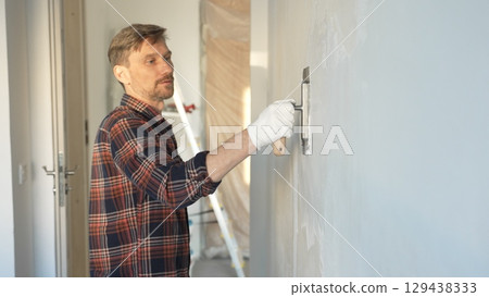 Male construction worker wearing protective gloves smoothing plaster onto wall surface during home renovation using professional trowel tool 129438333