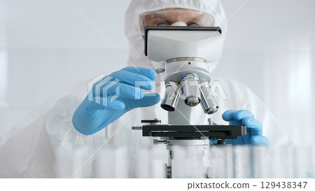Male scientific researcher or virologist wearing protective gear adjusting microscope, analyzing lab samples with precision in sterile research environment. Medicine concept Male scientific researcher or virologist wearing protective gear adjusting microscope, analyzing lab samples with precision in sterile research environment. Medicine concept 129438347