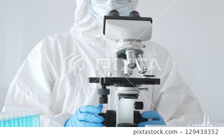 A male doctor in a protective suit and gloves uses a microscope while analyzing samples, conducting research in a laboratory. Close-up of test tubes with blue liquid 129438352
